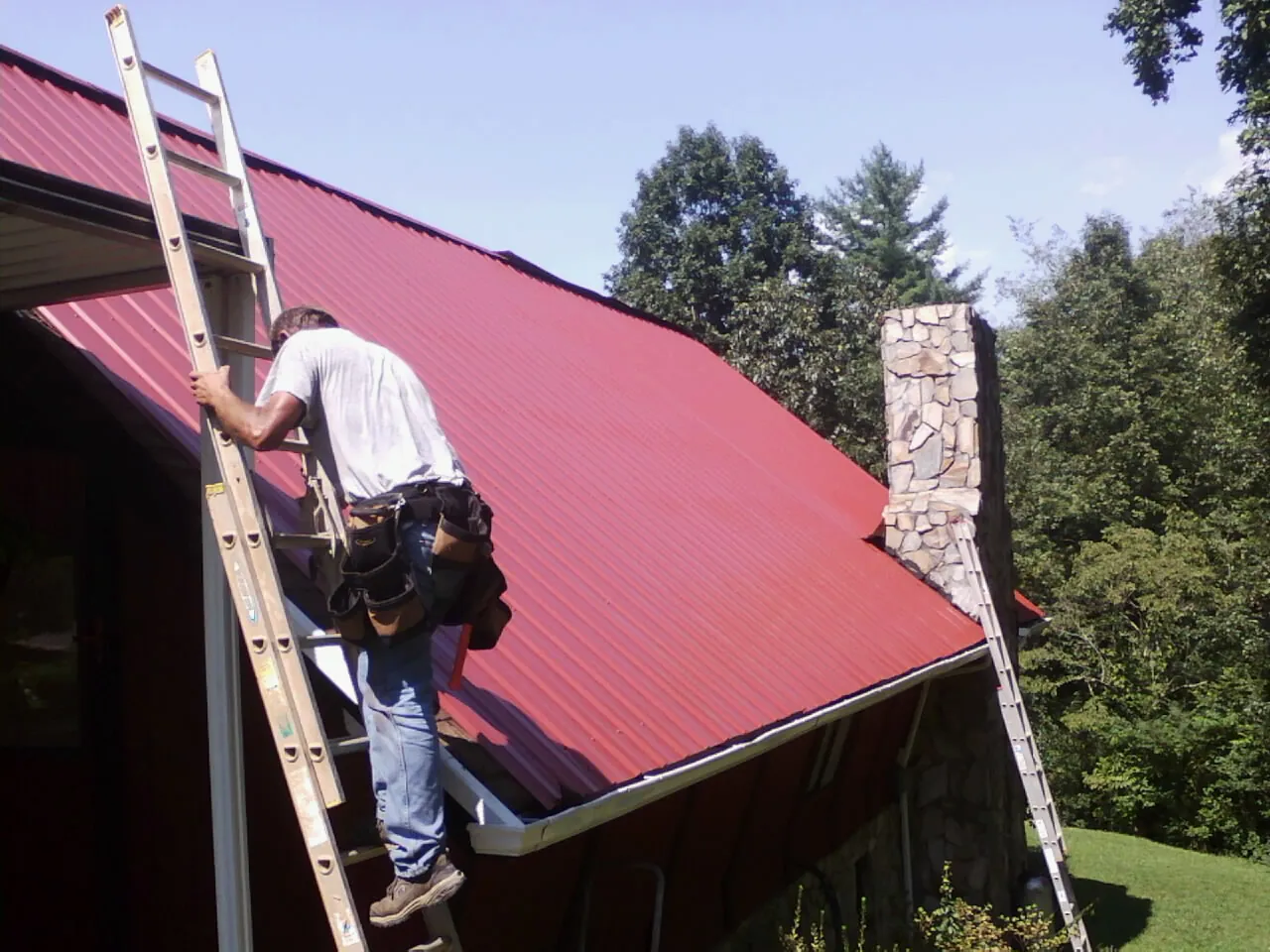 Roofer on a ladder beside a red metal roof and stone chimney — Soffit & Fascia Repair by Roofing Brothers in Mc Clellandtown